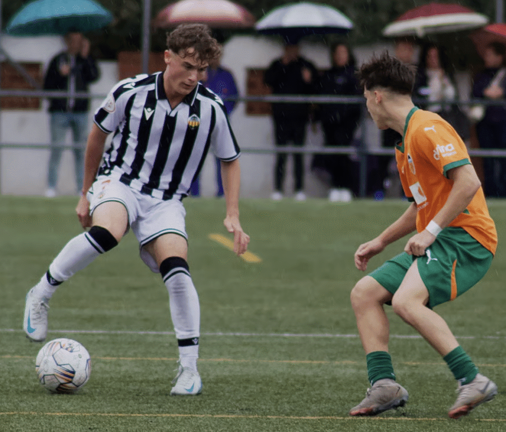 Jugador juvenil con camiseta blanquinegra encarando a rival durante un partido de fútbol en campo